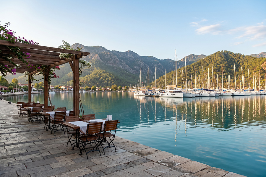 Göcek marina in autumn: white sailing yachts moored in glassy water, pine-covered mountains, and empty waterfront café tables. Once the summer crowds thin out, the town settles into a calm pace made for long working days — ideal for a workation. Info & booking: gocekrentals.com