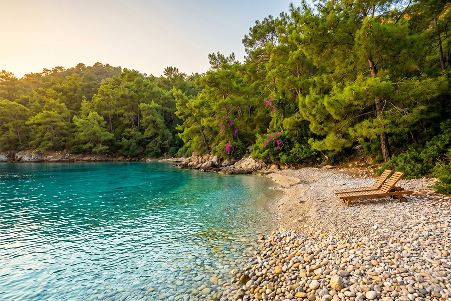 A quiet cove near Göcek: clear turquoise water, pine trees, and a single wooden lounger on the pebble shore. In October the sea is often still 23–24°C — perfect for a swim in the afternoon after a call. Info & booking: gocekrentals.com