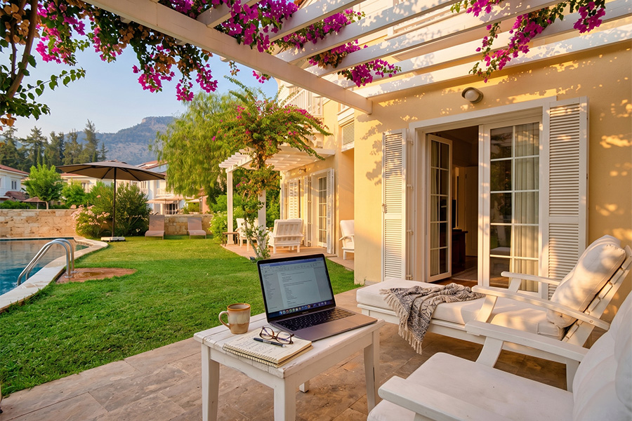 An open-air office on the terrace of our Portville villa, under a white bougainvillea-draped pergola: an open laptop, Turkish coffee and a notebook, with the pool and pine-covered hills behind. A calm workspace for autumn remote work in Göcek. Info & booking: gocekrentals.com